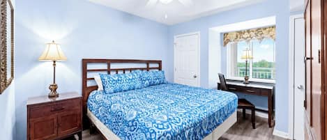 A bedroom with a bed, bedside table, lamp, desk, chair, and a window with a view.