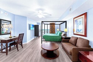 Ein Hotelzimmer mit Bett, Schreibtisch, Sessel, Sofa, Fernseher und Deckenventilator.