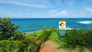 Camino costero con un cartel de advertencia por oleaje alto y precaución sobre zonas naturales peligrosas.