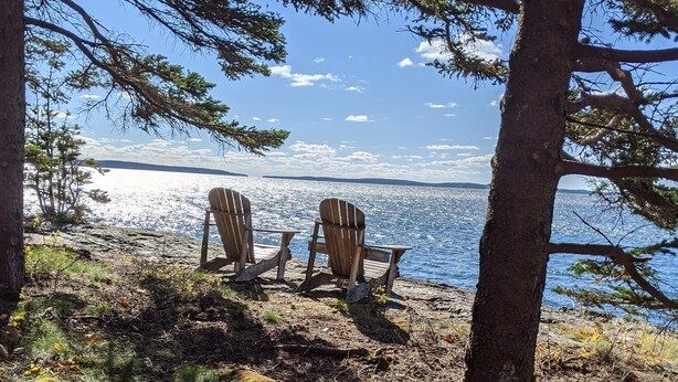 Two Adirondack chairs on a rocky shore with a view of a calm lake and distant mountains under a clear sky.