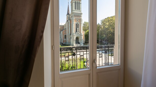 Blick durch ein Fenster auf eine Kirche mit einem Glockenturm, einen Balkon mit schmiedeeisernem Geländer und eine grüne Wiese.