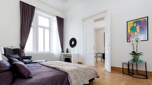 Ein Schlafzimmer mit Bett, Schminktisch, Fenster mit Vorhängen, Tür und einem Wandbild.
