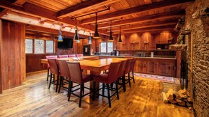 A spacious wooden interior with a large dining table, multiple chairs, pendant lights, a kitchen area, and a stone wall.