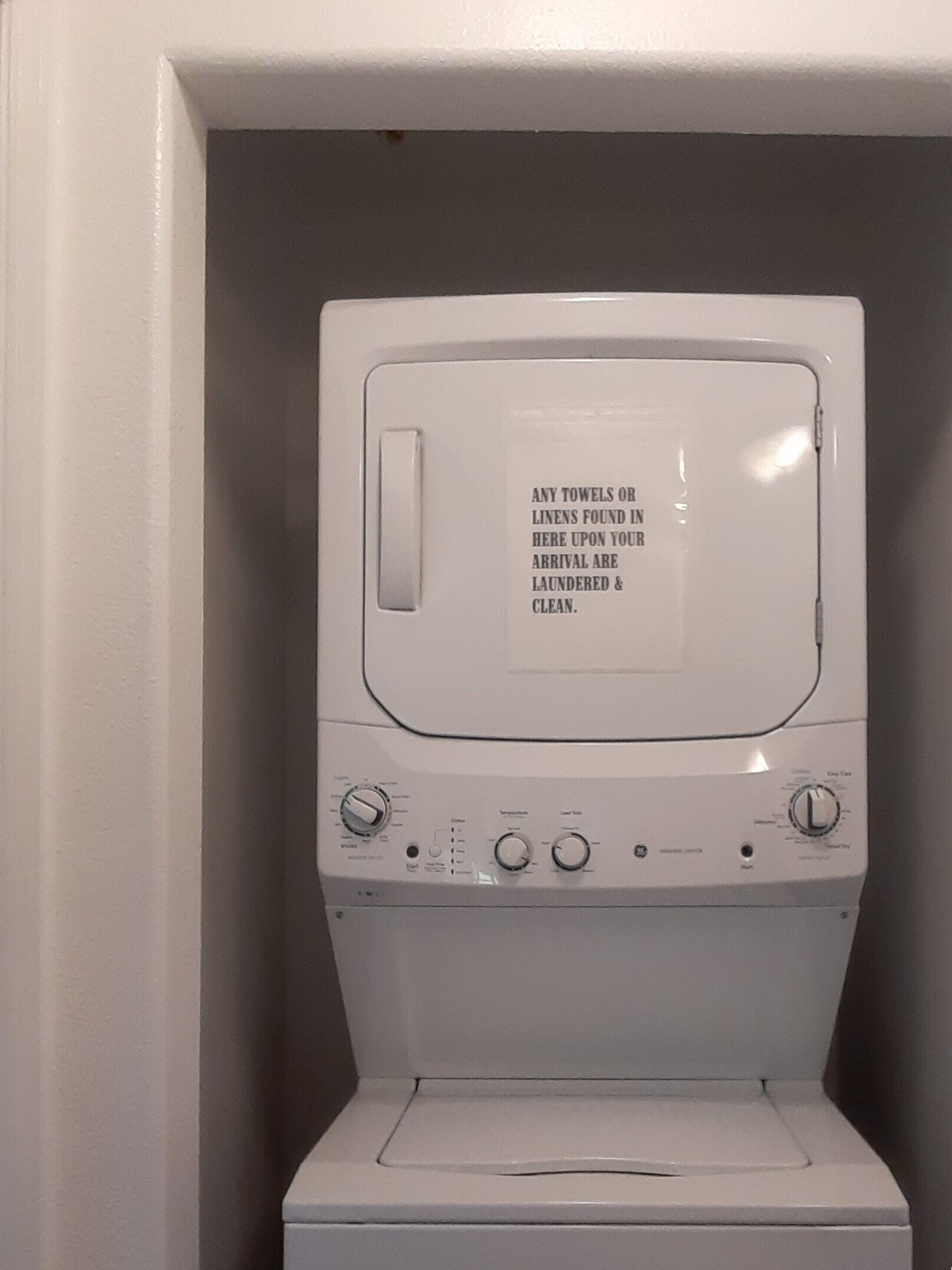 Una lavadora blanca con un mensaje en la puerta que indica que cualquier toalla o ropa de cama que se encuentre dentro al llegar, ha sido lavada y está limpia.