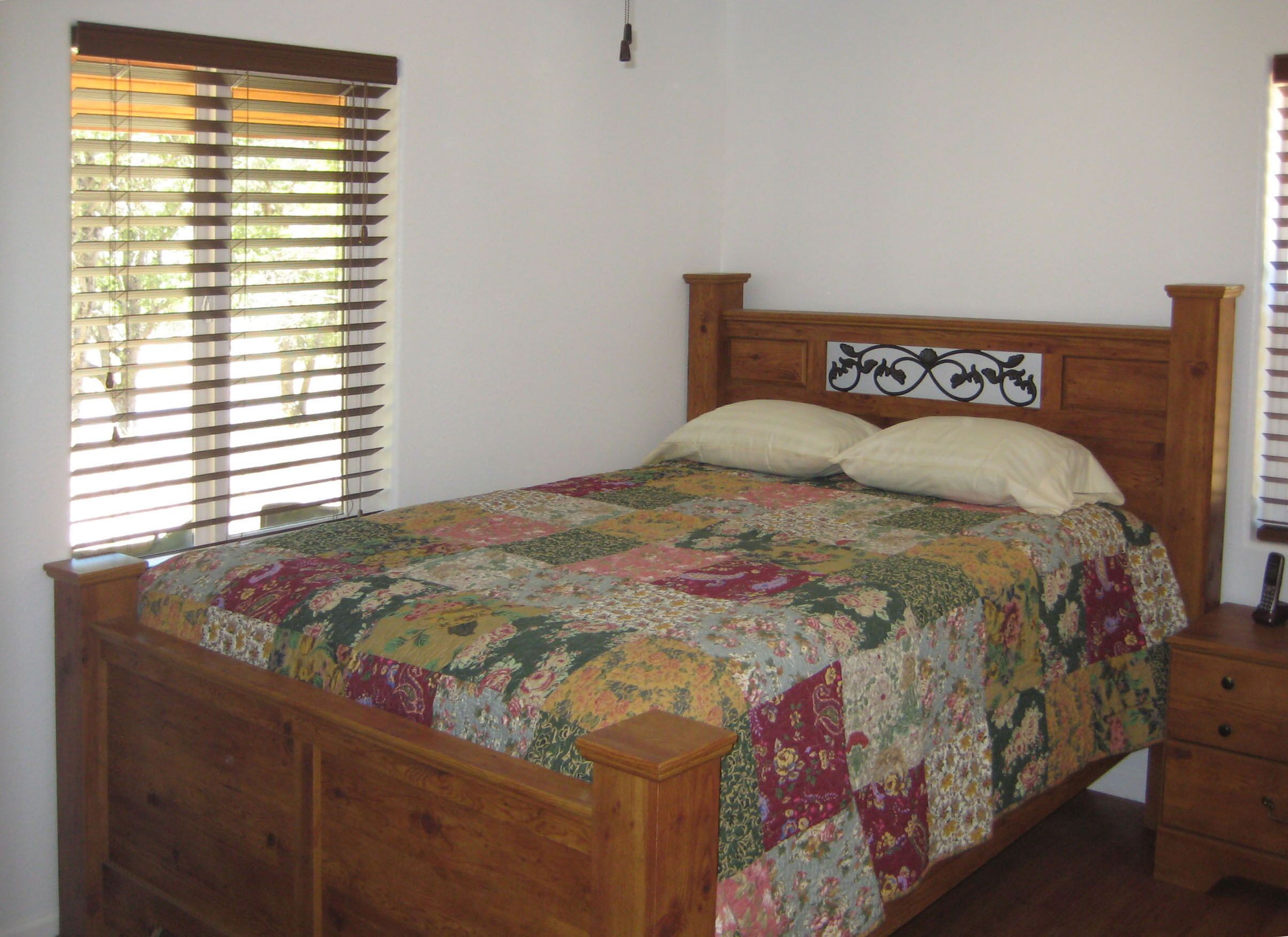 Uma cama de madeira com colcha florida, uma mesinha de cabeceira e uma janela com persianas.