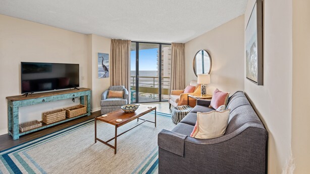 A modern living room with a flat-screen TV, a wooden coffee table, a grey sofa, and a large window offering a view of the city. - Oceanfront condo in private community + Free Attraction Tickets! (Myrtle Beach)