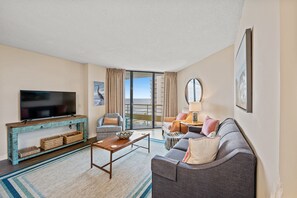 A modern living room with a flat-screen TV, a wooden coffee table, a grey sofa, and a large window offering a view of the city. - Oceanfront condo in private community + Free Attraction Tickets! (Myrtle Beach)