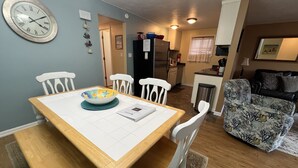 Uma área de refeições com uma mesa de madeira e cadeiras brancas, uma cozinha ao fundo e uma poltrona estampada.