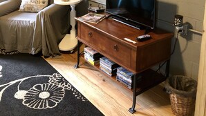 A cozy living room with a television, a wooden cabinet with books, a pet-themed pillow, and a welcome sign on the wall.