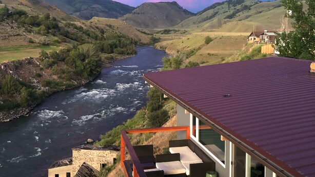 A modern house with a deck overlooking a river and hills.