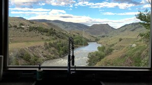 A window view of a river valley with mountains, a metal structure in the foreground, and a bottle of hand sanitizer on the windowsill.