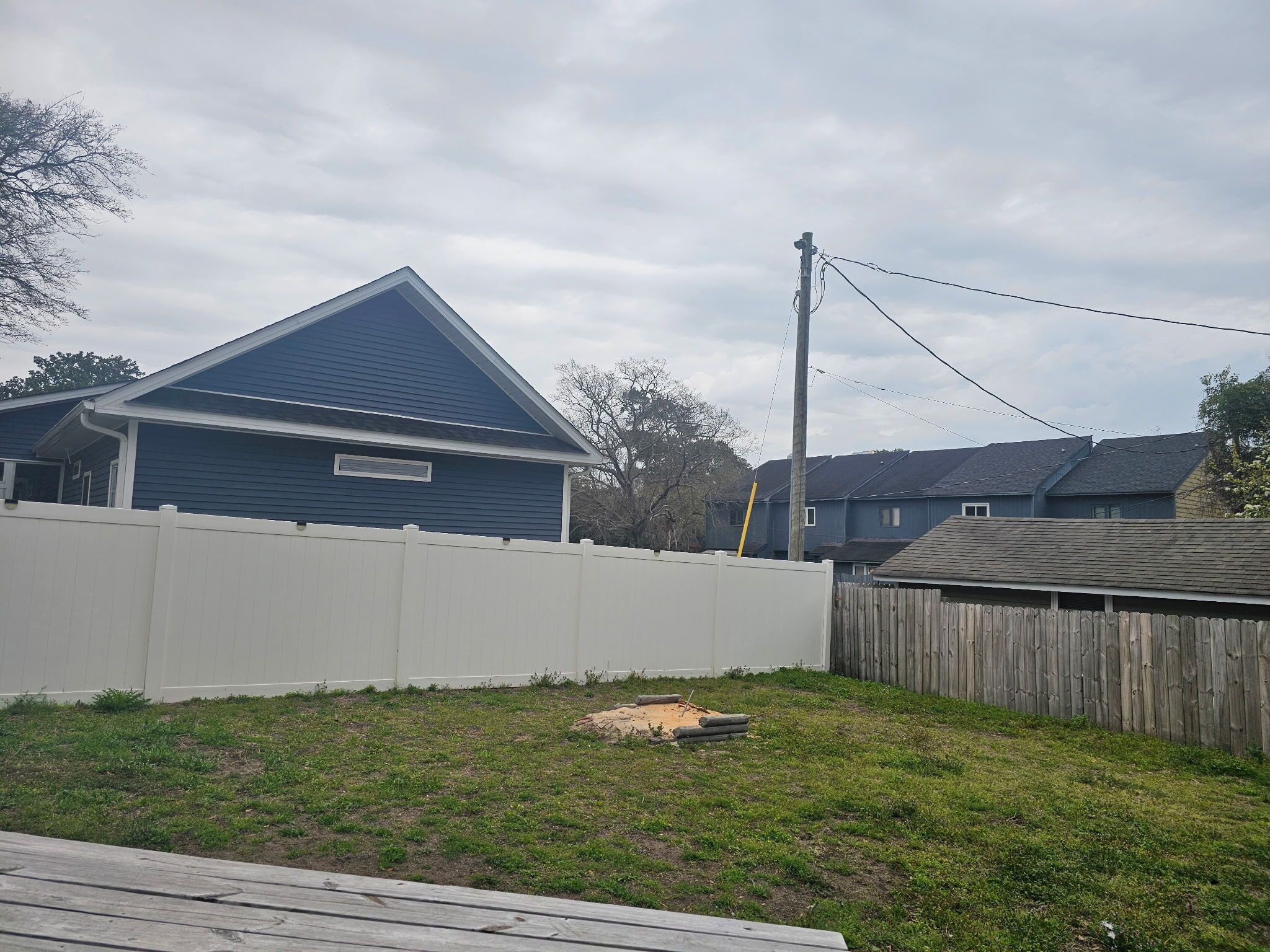 A residential backyard with a white fence, a wooden deck, and a blue house with a gable roof.