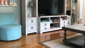 A living room with a wooden floor, a blue ottoman, a TV stand with drawers, and a painting on the wall. - Fully Fenced in Backyard, 1 block to the beach, Golf Cart included, (Myrtle Beach)