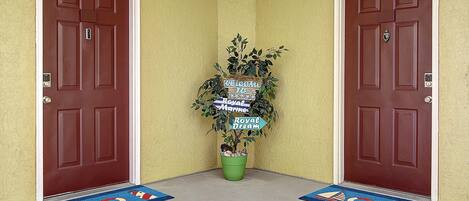 Een hoekje met twee bruine deuren, een welkomstbord en een plant in pot.