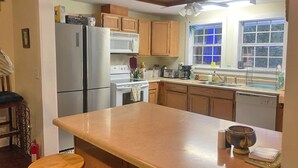 Ett kök med träskåp, ett kylskåp i rostfritt stål, en mikrovågsugn, en spis och en diskmaskin. Det finns ett fönster med en blå gardin, en träpall och en kaffebryggare på bänkskivan.