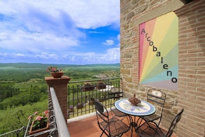 Un balcon donnant sur un paysage verdoyant, une table avec des chaises et une enseigne colorée sur le mur.