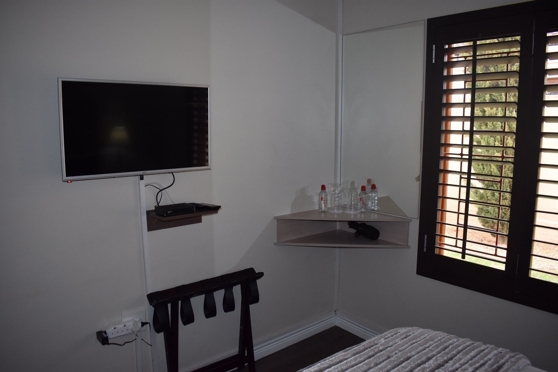 A bedroom with a TV mounted on the wall, a window with blinds, a bedside table with a lamp, and a bed with a blanket.