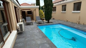 Una piscina con baldosas grises, un porche cubierto con sillones de descanso y un edificio de fachada color crema.