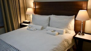 Una cama bien tendida con sábanas blancas y dos toallas enrolladas, un cabecero de madera y dos lámparas de noche.
