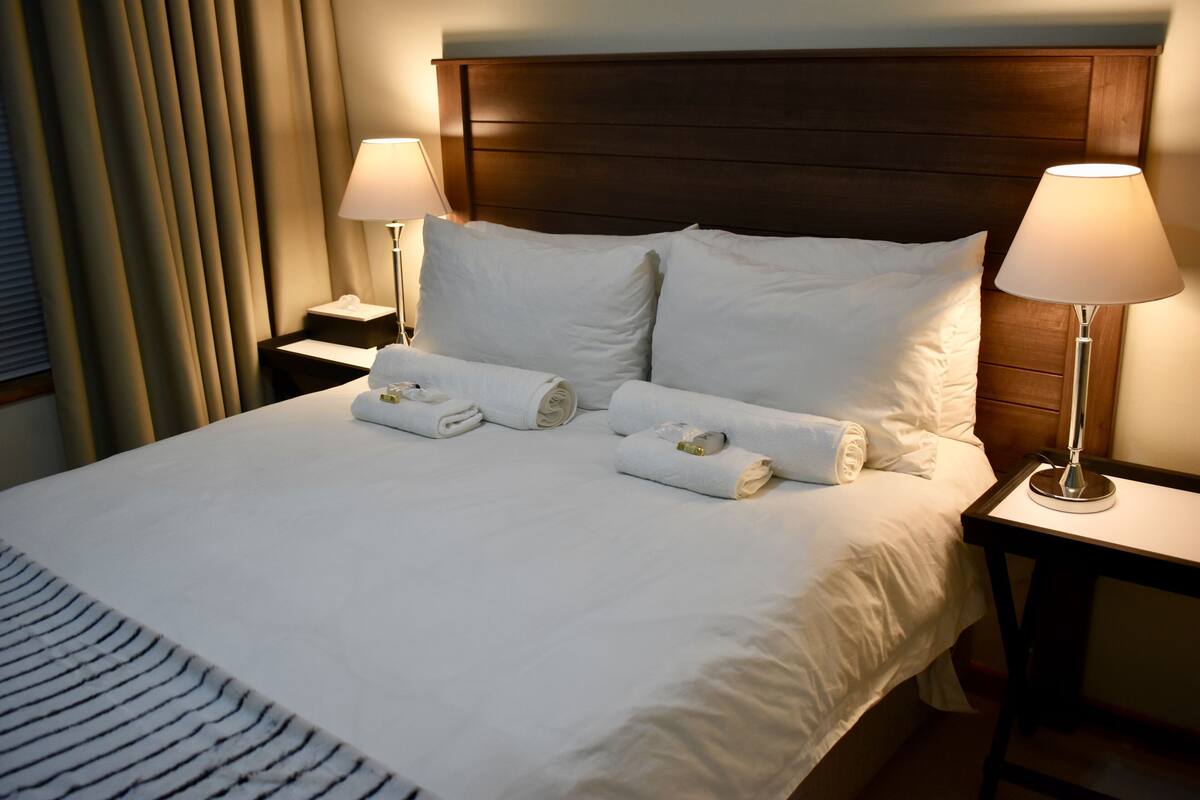 A neatly made bed with white linens and two rolled towels, a wooden headboard, and two bedside lamps.