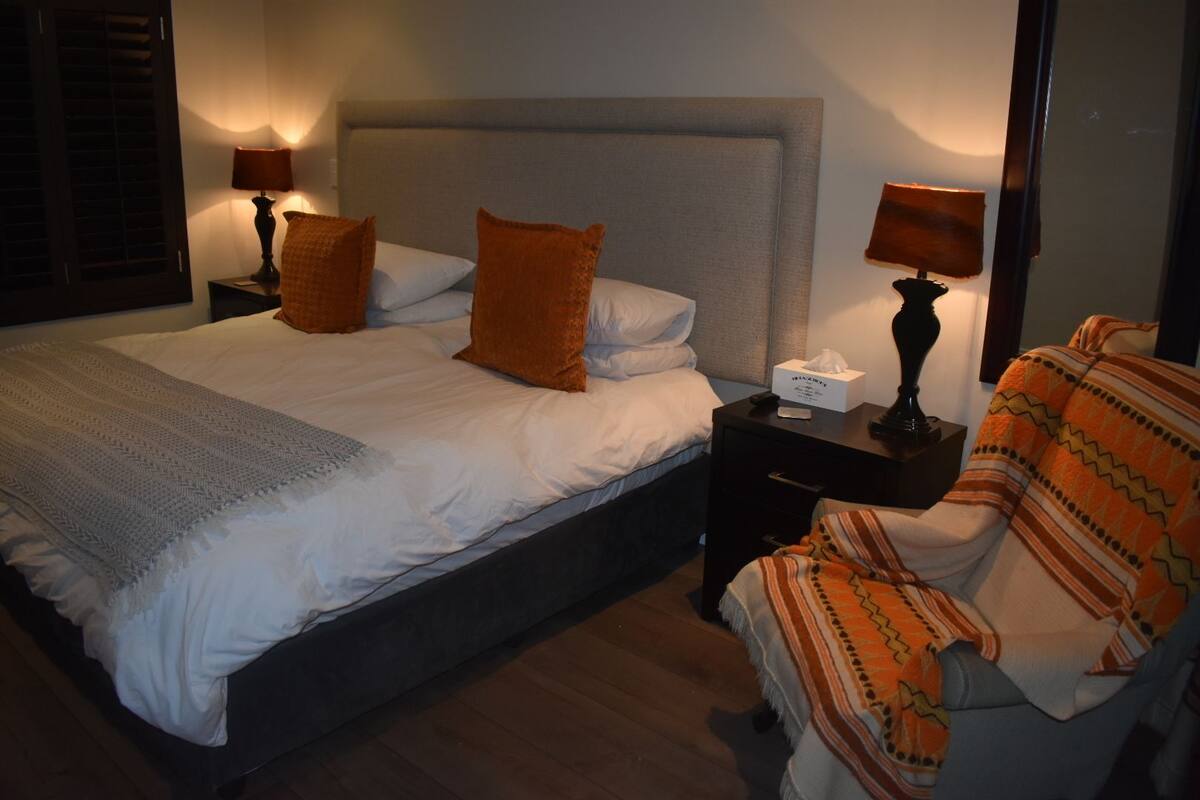 A hotel room with a large bed, two bedside tables with lamps, a chair with a blanket, and a bedside box of tissues.