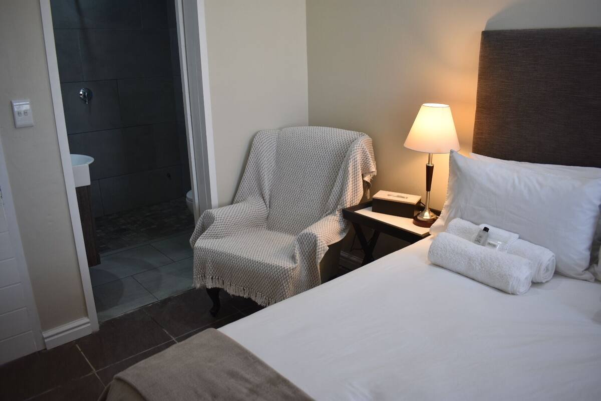 A hotel room with a bed, a chair, a nightstand with a lamp, and a shower area.