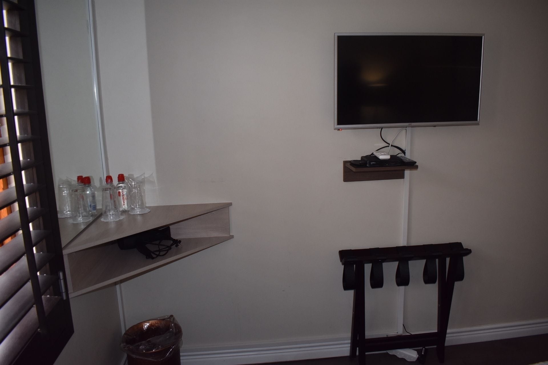 Una habitación con una televisión montada, un estante con botellas de agua, un cubo de basura y una silla.