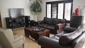 Una sala de estar con un sofá de cuero marrón, un sillón beige, una mesita de centro, un televisor y altavoces.
