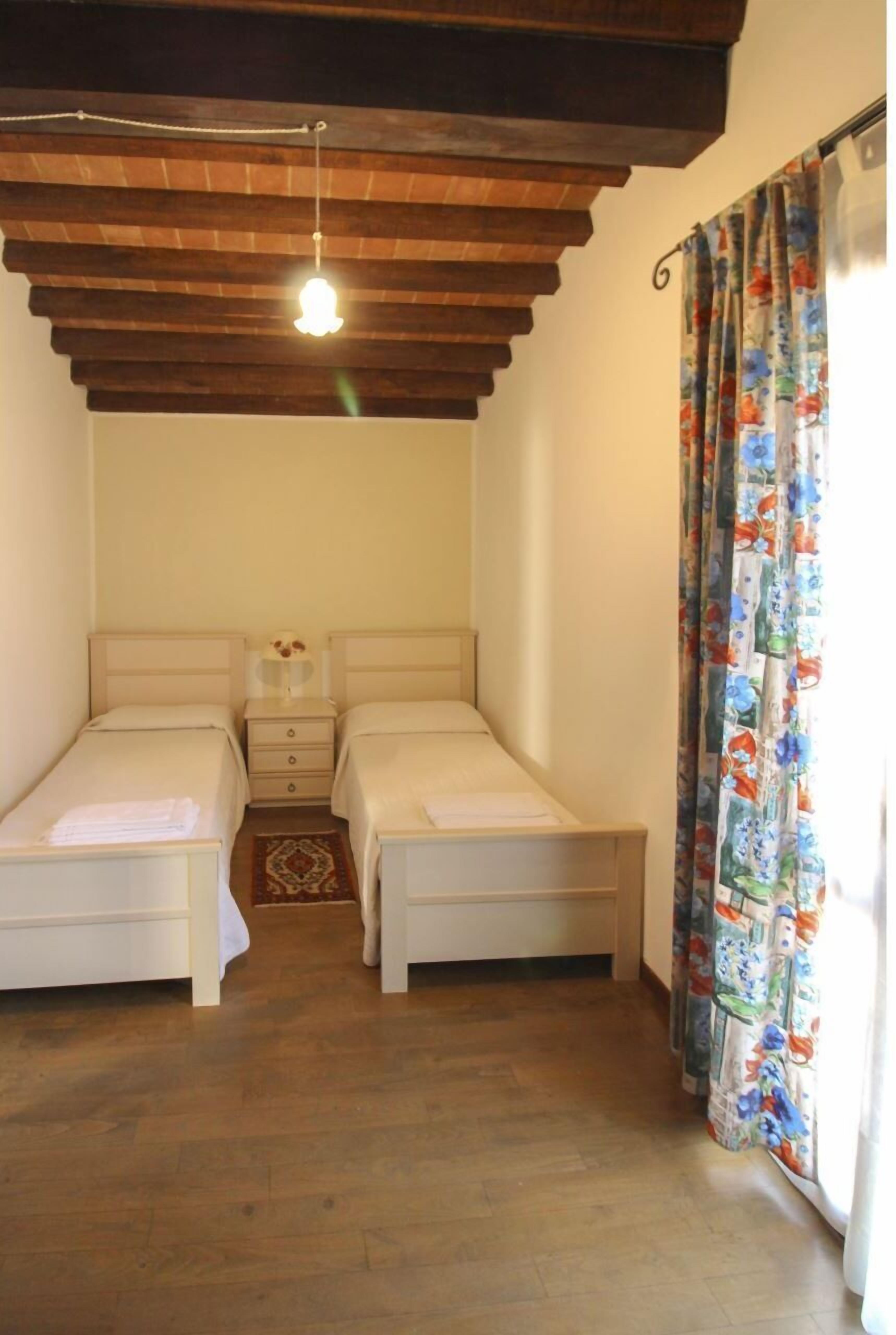Un dormitorio con dos camas, techo de madera, mesita de noche y cortina estampada.
