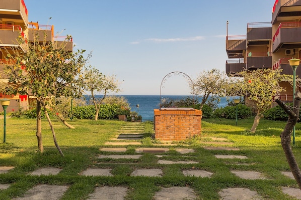 A pathway leading to a brick wall with a metal archway, surrounded by greenery and buildings with balconies overlooking a body of water.