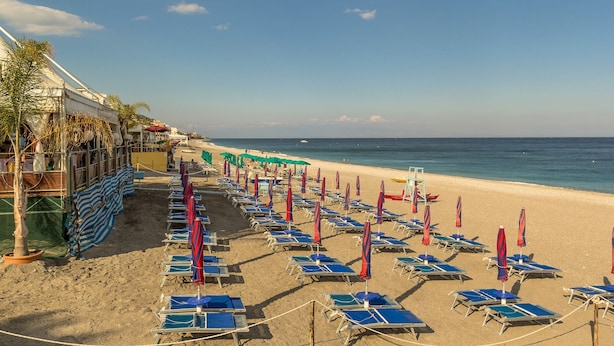 A beach with rows of blue lounge chairs and red umbrellas, a sandy shore, and a clear blue sky. - Blue Wood Apartment (Letojanni)