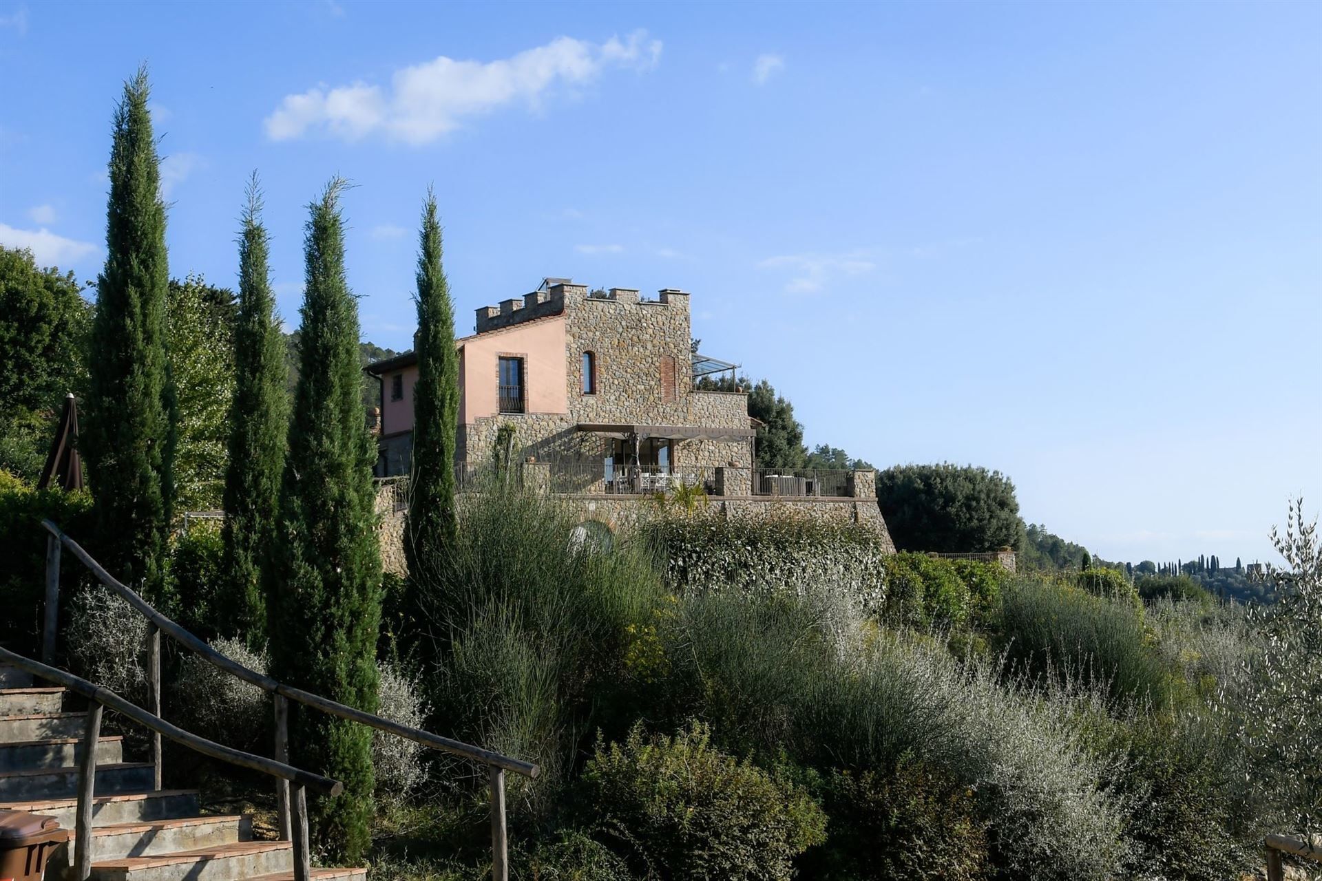 Un bâtiment en pierre à la façade rose, entouré de grands cyprès et d’une végétation dense.