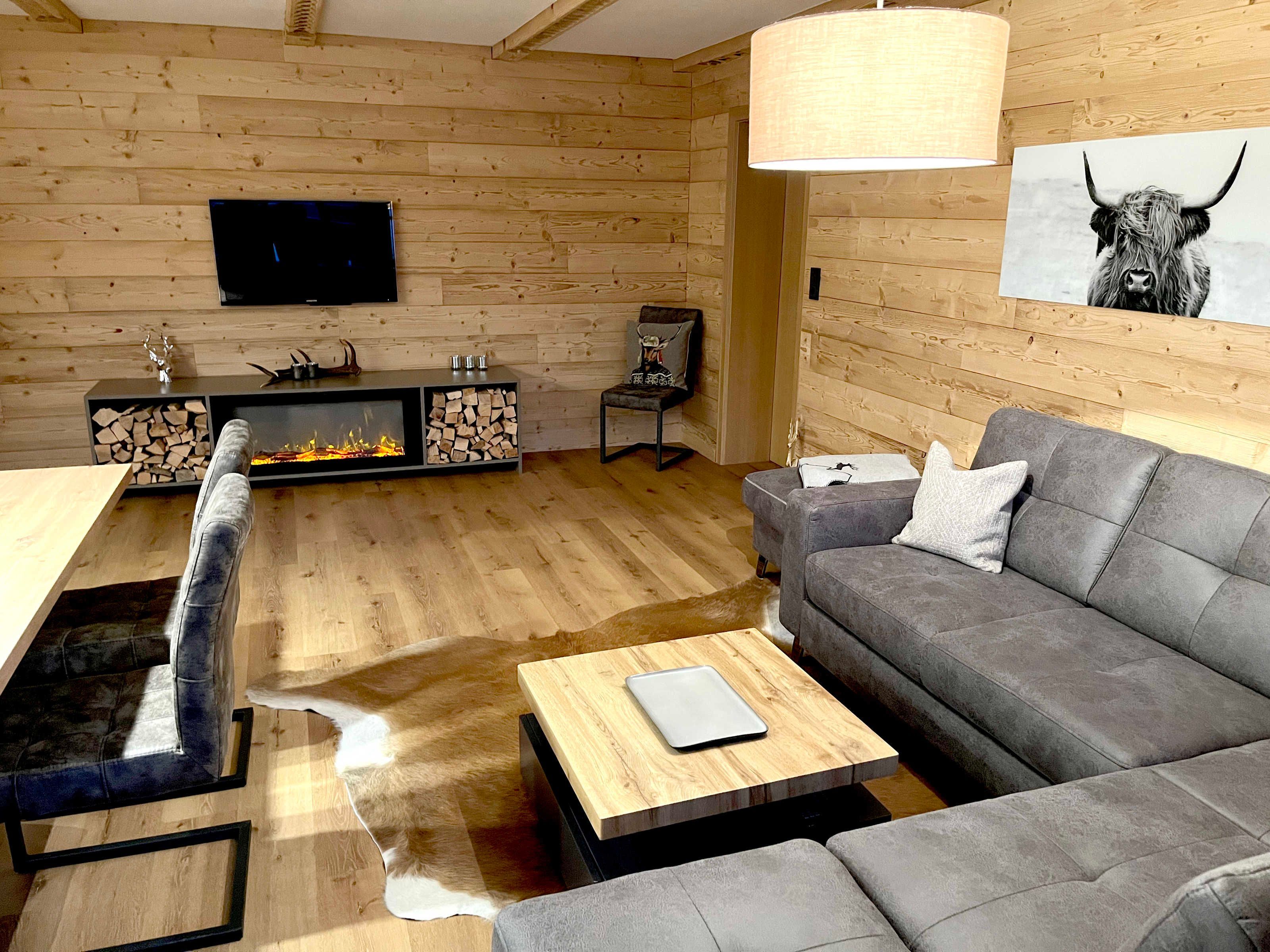 Un salon moderne avec une cheminée, un canapé gris, une table basse en bois et un téléviseur fixé au mur.