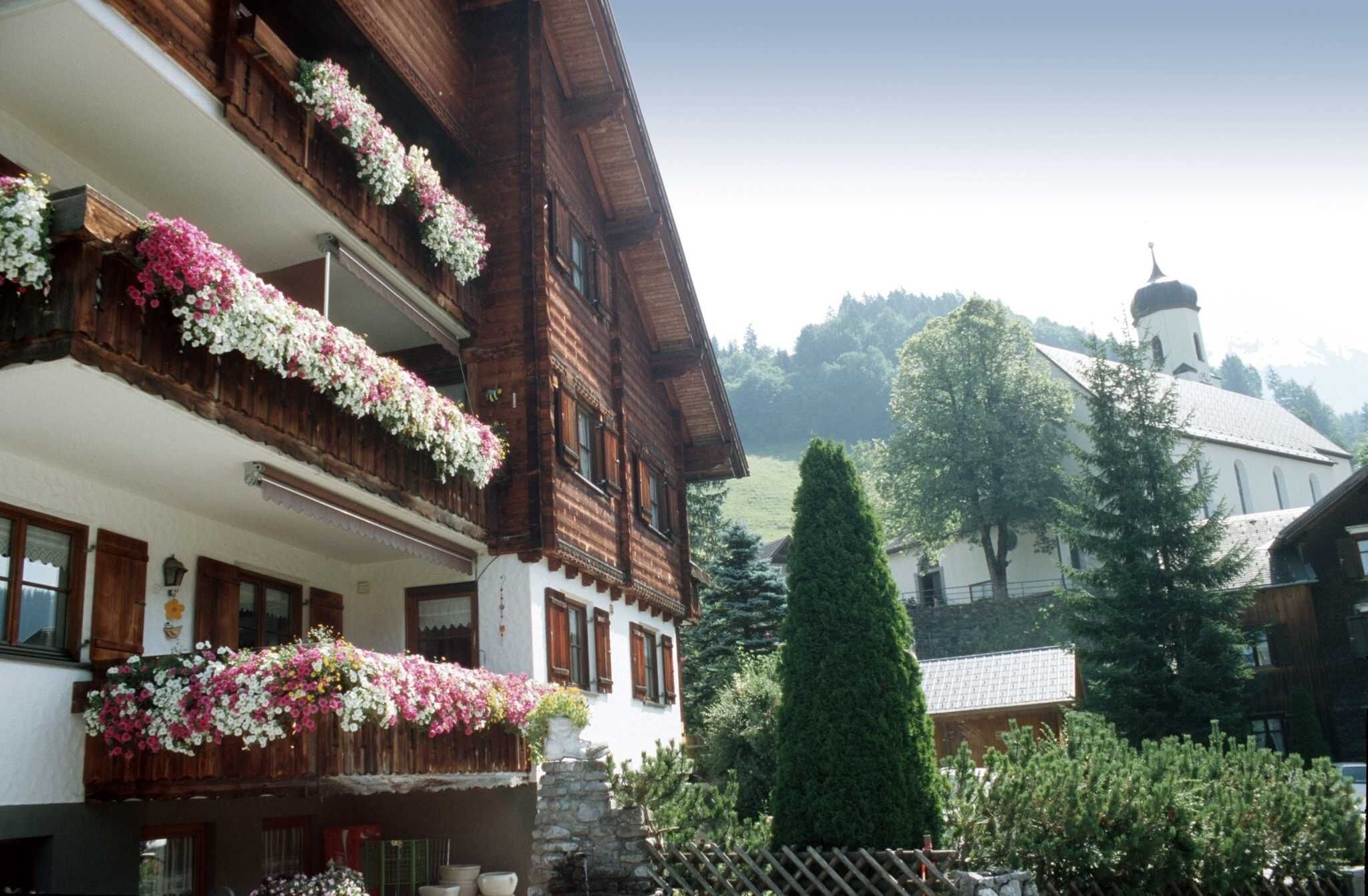 Un edificio in legno tradizionale con balconi decorati da fiori, una chiesa sullo sfondo e un giardino curato.