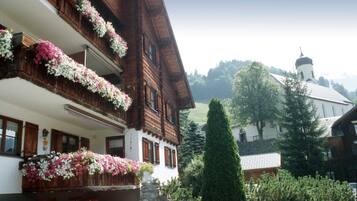 Un edificio in legno tradizionale con balconi decorati da fiori, una chiesa sullo sfondo e un giardino curato.