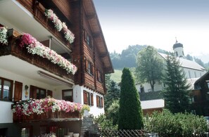 Ein traditionelles Holzhaus mit Balkonen, die mit Blumen geschmückt sind, sowie eine Kirche im Hintergrund und ein gepflegter Garten.