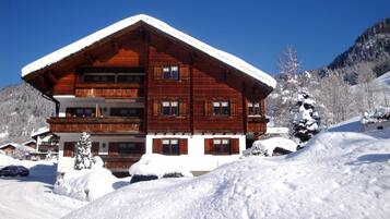 A multi-story wooden chalet with a snow-covered roof, surrounded by snow-covered trees and a mountainous backdrop.