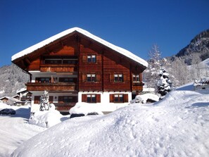 Una cabaña de madera de varios pisos con techo nevado, rodeada de árboles cubiertos de nieve y un paisaje montañoso.