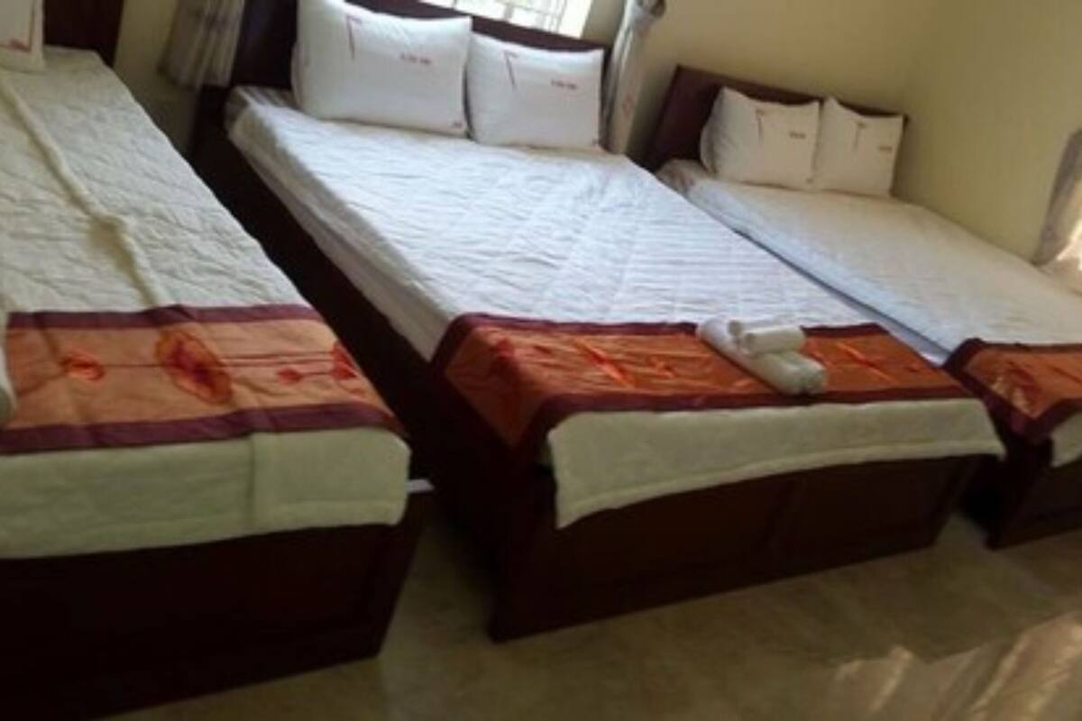 A room with three single beds, each with a white mattress and a brown headboard. The beds are neatly made with white sheets and orange blankets. There are pillows on each bed, and towels are folded on the beds.