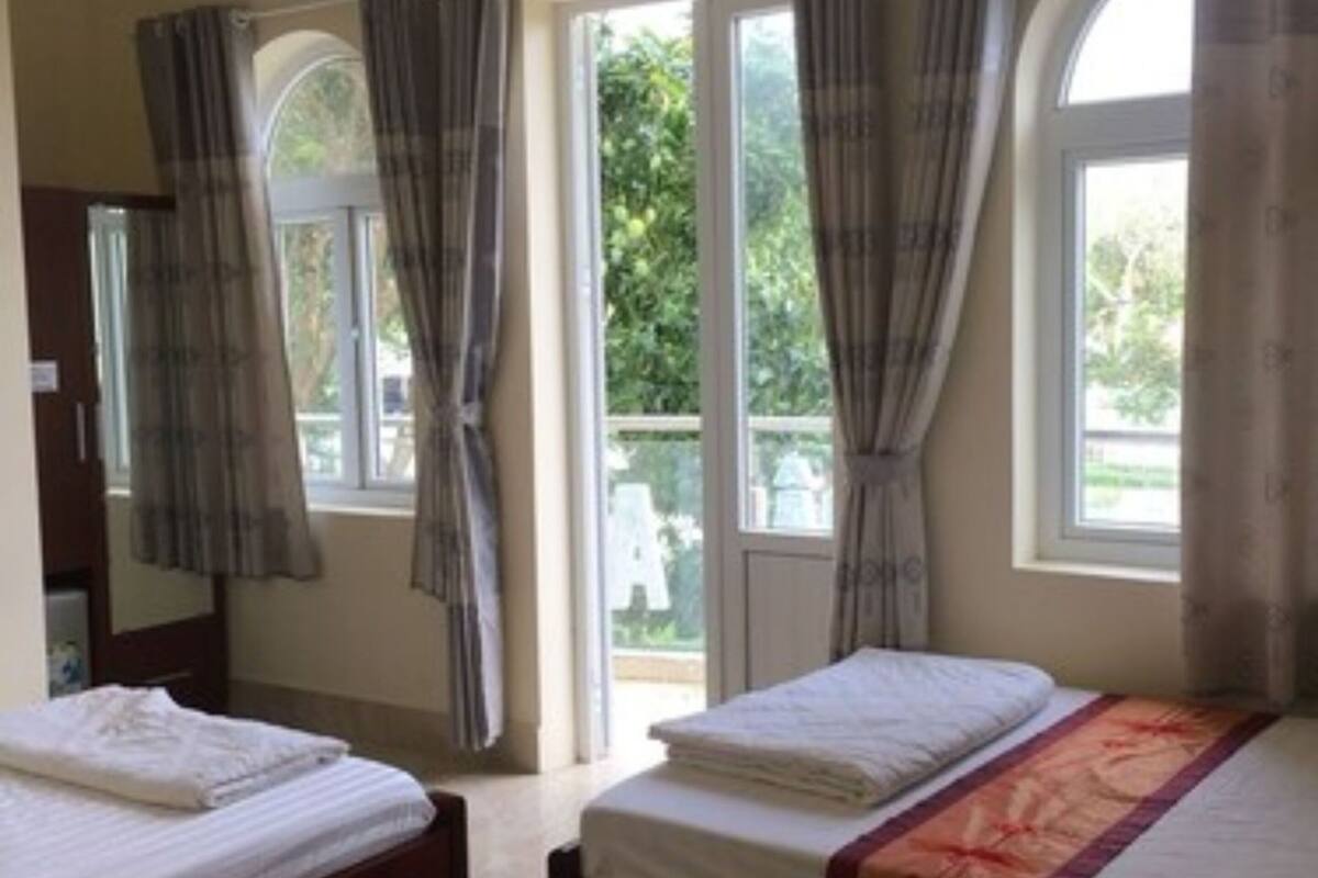 A room with two beds, a large window with curtains, and a view of greenery outside.