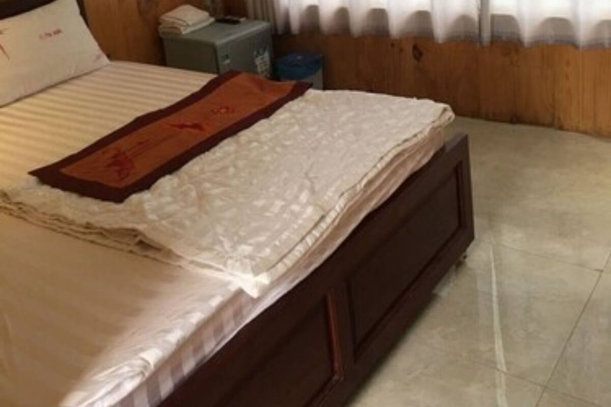 A bedroom with a wooden headboard, a bed with a white comforter and a brown throw, a bedside table with a lamp, and a window with sheer curtains.