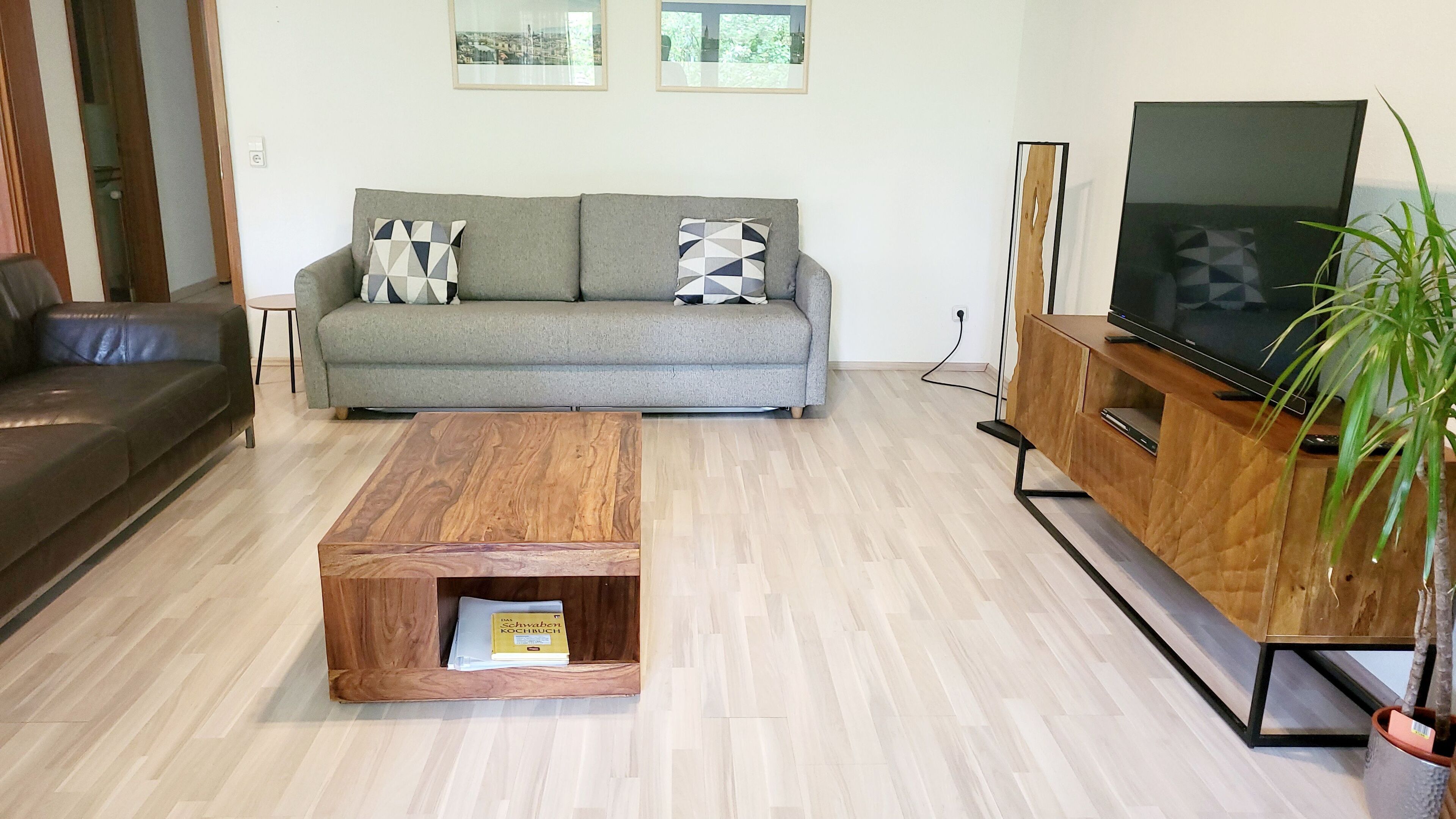 Ein Wohnzimmer mit grauem Sofa, einem Holztisch, einem Fernseher auf einem Holzschrank und einer Topfpflanze.