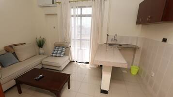 Un espace de vie compact comprenant un canapé, une petite table basse, une kitchenette avec un évier et un plan de travail, et une fenêtre avec des rideaux.