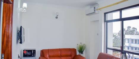 A compact living space with a brown leather sofa, a small kitchenette with a microwave, and a window providing a view of buildings outside.