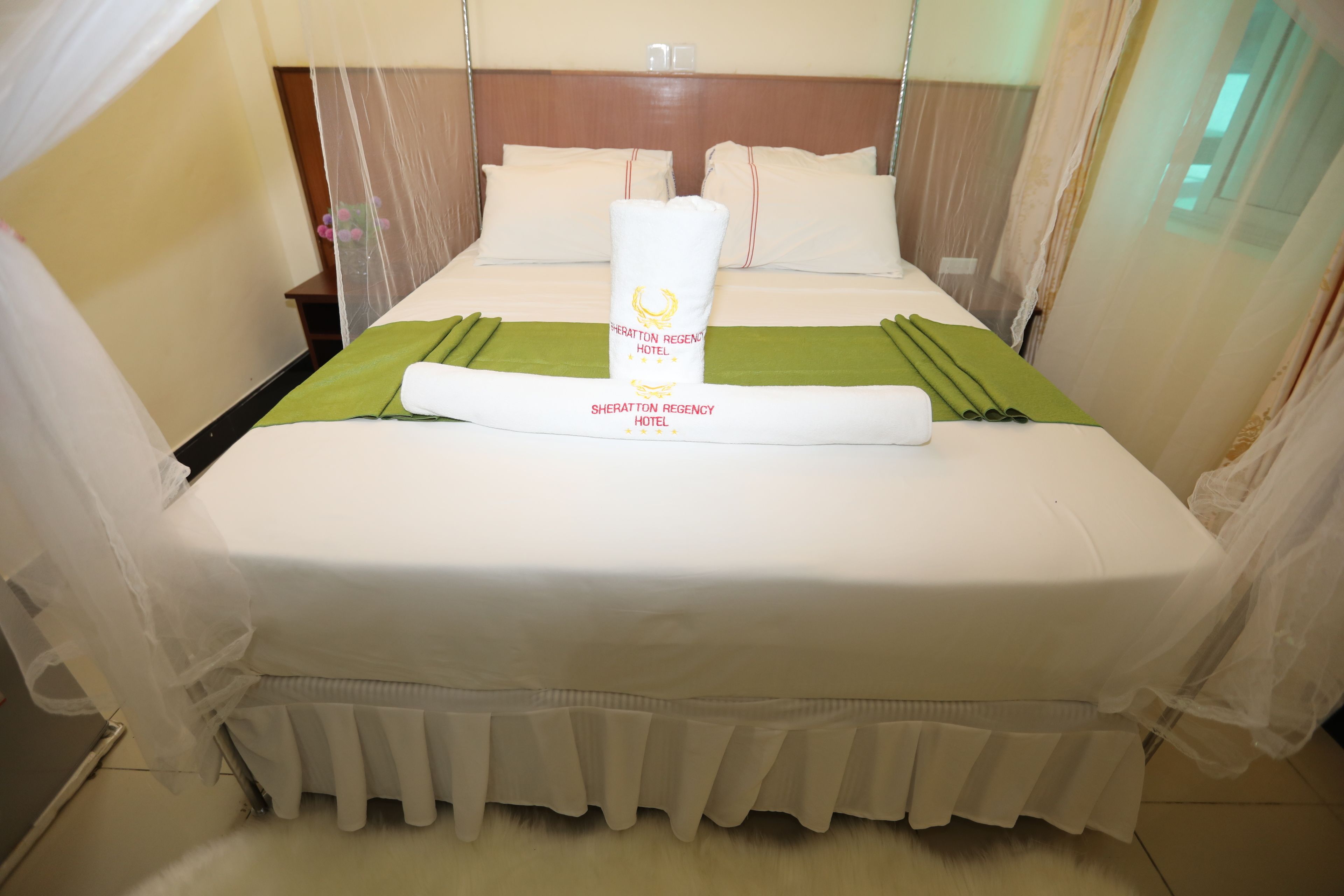 Een netjes opgemaakt bed met een groene sprei en een opgerolde handdoek in het midden, een houten hoofdbord en dunne gordijnen voor de ramen.