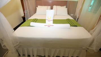 Una cama bien hecha con una colcha verde y una toalla enrollada en el centro, un cabecero de madera y cortinas translúcidas en las ventanas.