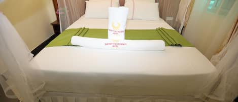 A neatly made bed with a green bedspread and a rolled towel in the center, a wooden headboard, and sheer curtains on the windows.