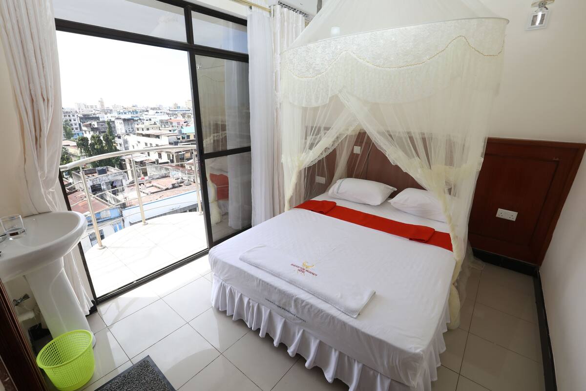 A hotel room with a balcony, a bed with a canopy, a sink, and a green basket.