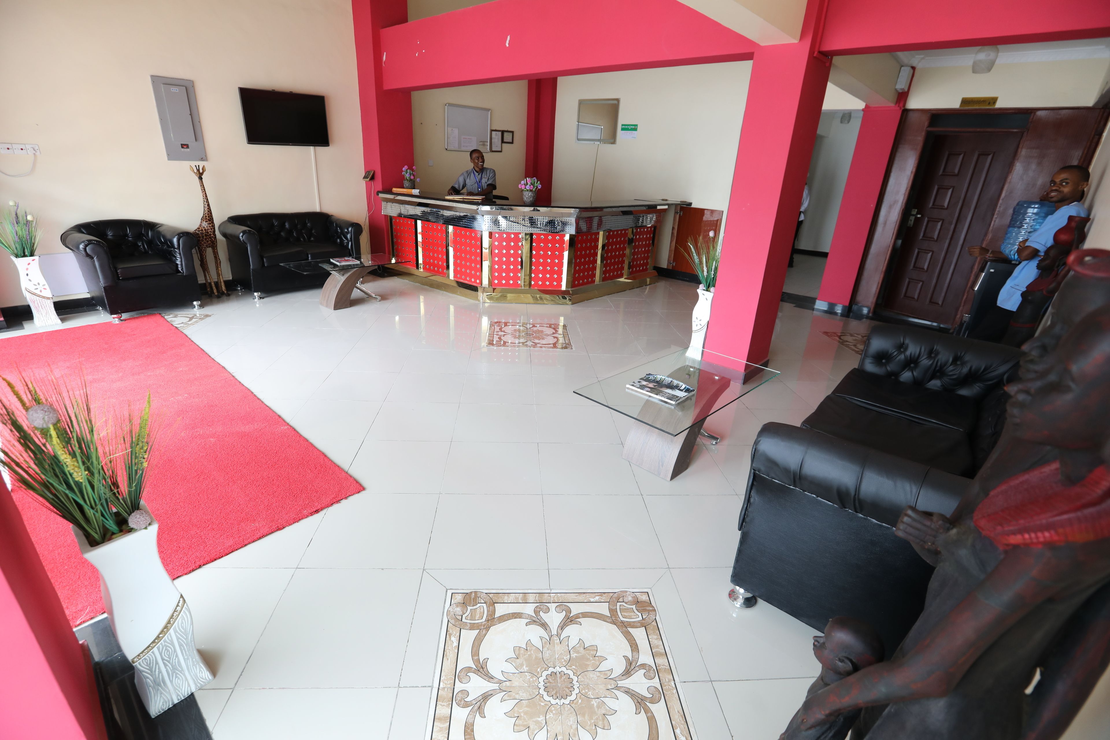 A hotel lobby with a reception desk, black leather sofas, a red carpet, and a decorative vase with plants.