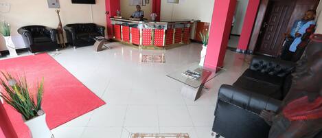 A hotel lobby with a reception desk, black leather sofas, a red carpet, and a decorative vase with plants.